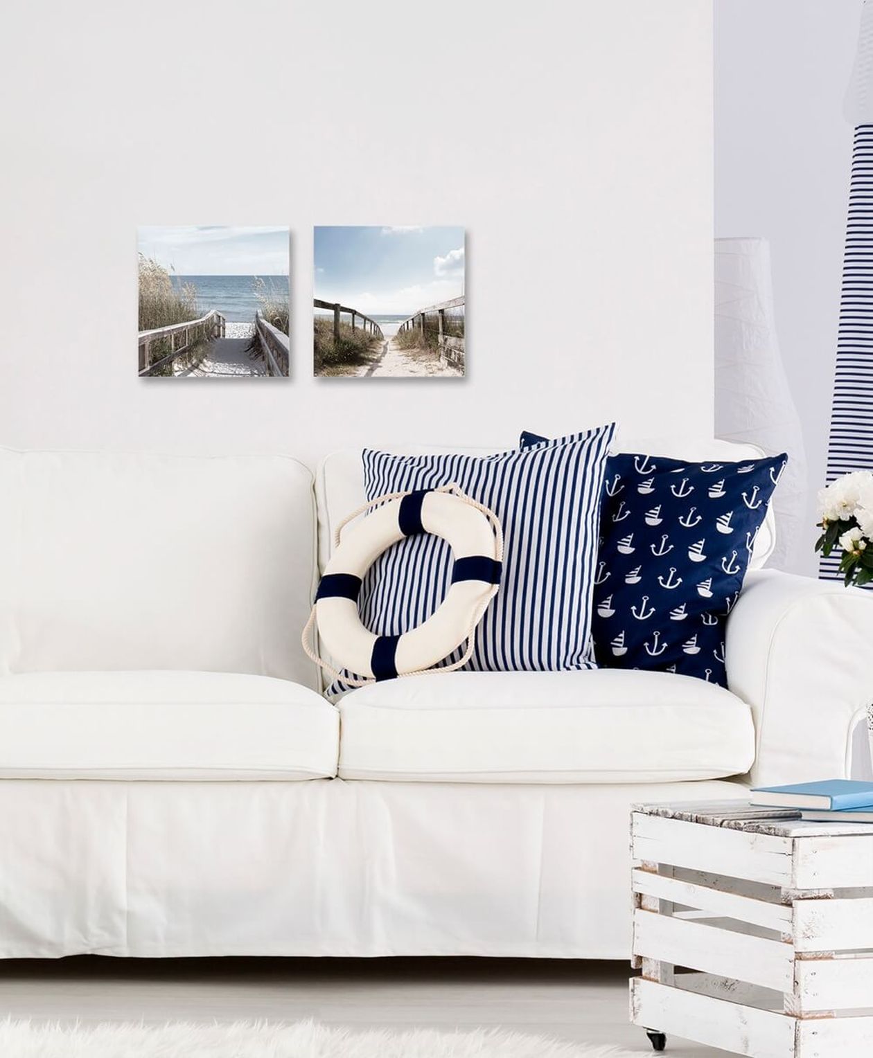Zwei quadratische Glasbilder mit Strandwegen an einer weißen Wand über einem weißen Sofa. Maritime Dekokissen mit Anker- und Streifenmuster sowie ein Rettungsring als Deko.