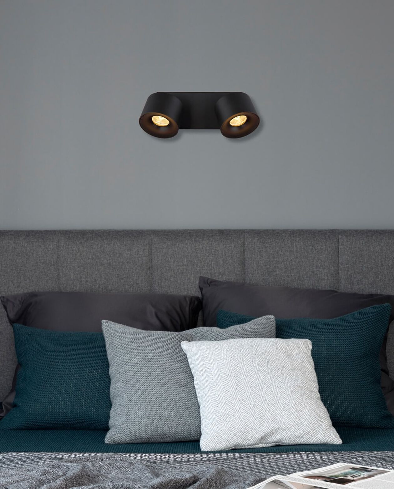 Schwarzer LED-Doppelstrahler ist an der Wand über einem Bett mit dunklen Kissen angebracht.