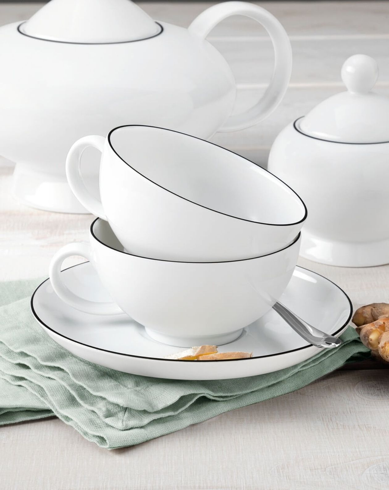 Zwei gestapelte, weiße Kaffeetassen mit dünnem schwarzen Rand und Untertasse, auf pastellgrüner Serviette