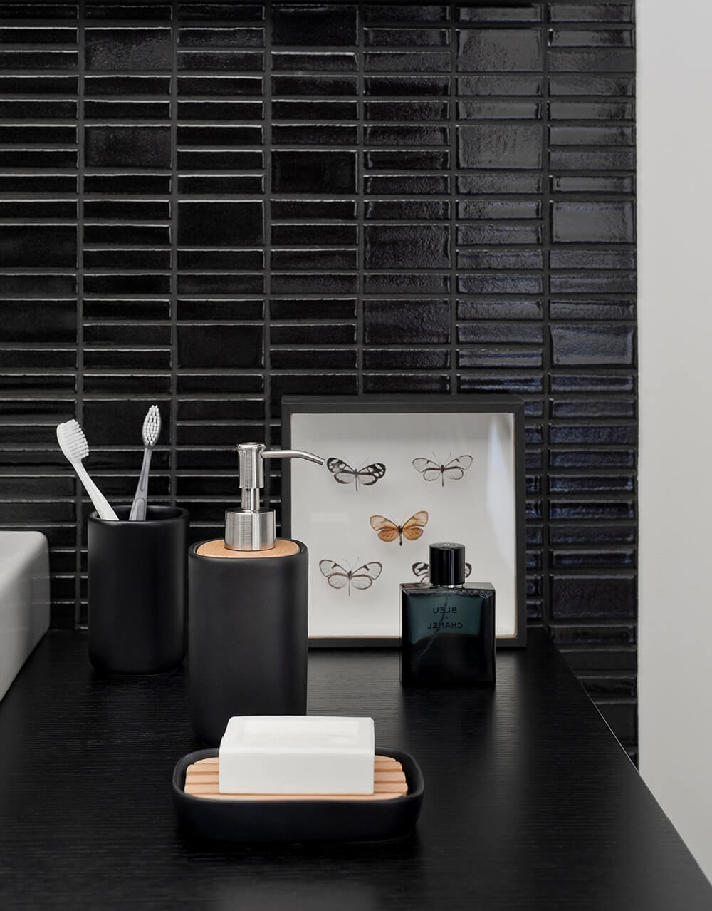Badezimmerarbeitsplatte mit schwarzen Accessoires, darunter ein Seifenspender, ein Zahnbürstenhalter, eine Seifenschale mit weißer Seife und ein gerahmtes Schmetterlingsbild.