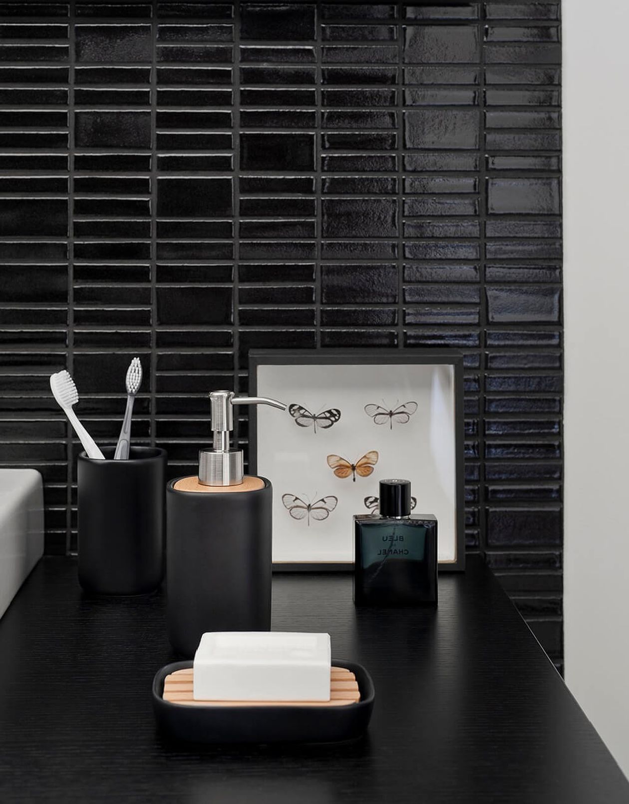 Badezimmerarbeitsplatte mit schwarzen Accessoires, darunter ein Seifenspender, ein Zahnbürstenhalter, eine Seifenschale mit weißer Seife und ein gerahmtes Schmetterlingsbild.