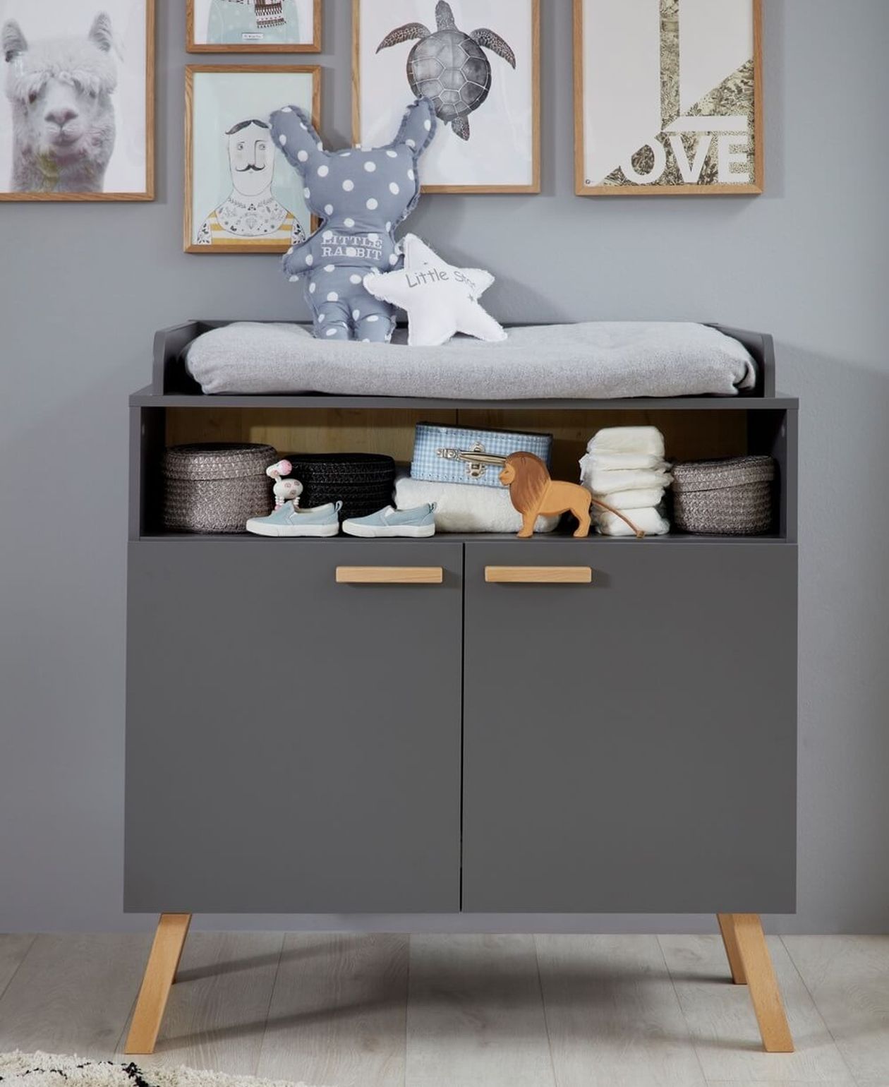 Graue Wickelkommode mit Holzgriffen und Wickelaufsatz, offenes Mittelfach mit Windeln und Spielzeug, auf zwei Holzfüßen, vor einer grauen Wand mit gerahmten Tierbildern.