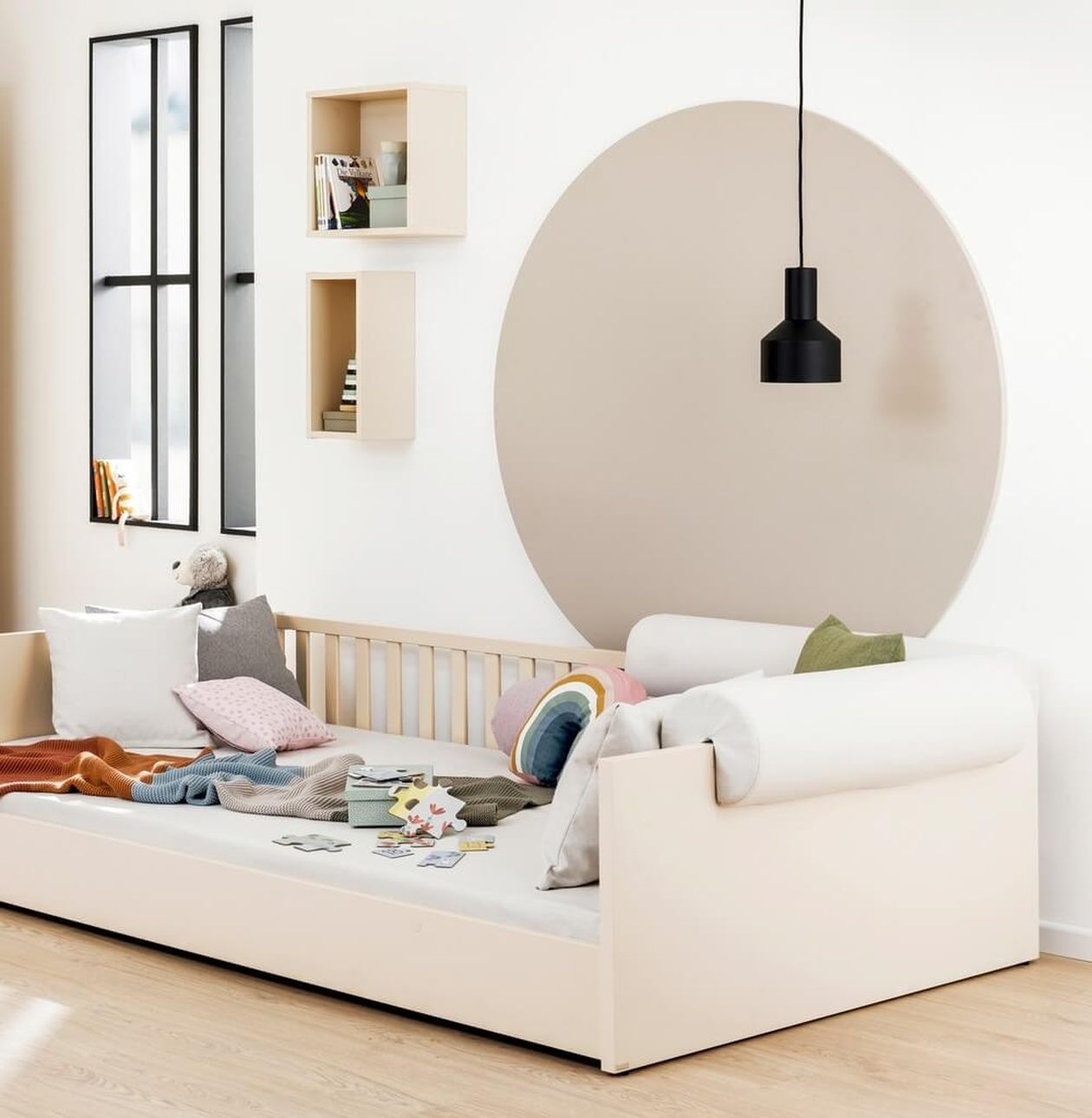 elles Kinderzimmer mit niedrigem weißem Kinderbett mit Holzsprossen, bunten Kissen und Spielzeug. An der Wand ein großer beigefarbener Kreis als Dekoration, darüber eine schwarze Pendelleuchte. Zwei quadratische Wandregale aus hellem Holz.