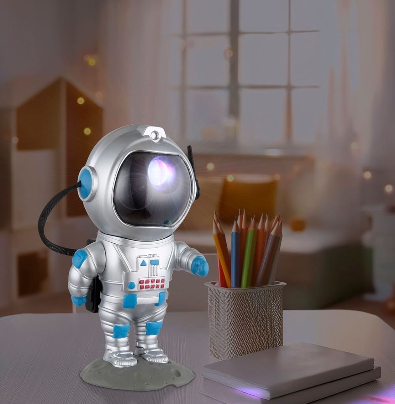 Silberne LED-Tischleuchte in Form eines Astronauten mit blauen Akzenten und leuchtendem Visier, die bunte Lichteffekte projiziert. Die Leuchte steht auf einem Schreibtisch neben Buntstiften und Heften in einem Kinderzimmer.