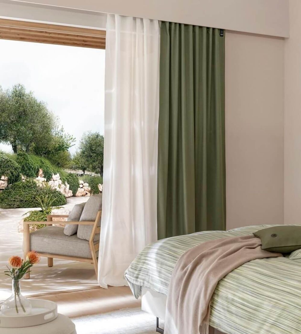 Helles Schlafzimmer mit Blick auf eine Terrasse, weiße transparente Gardine neben einem grünen Vorhang, Bett mit gestreifter Bettwäsche in Grüntönen.