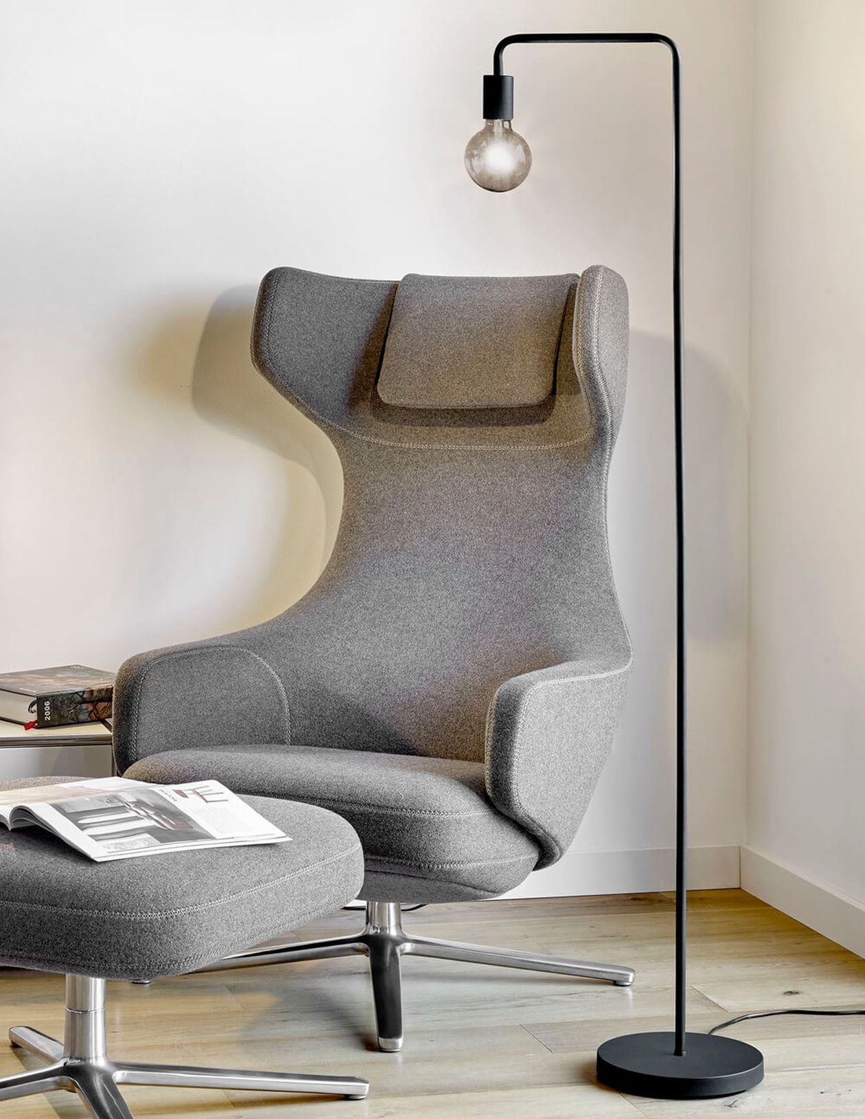 Ein moderner grauer Sessel mit passendem Hocker steht neben einer eleganten schwarzen Stehlampe und einem kleinen Tisch mit einem aufgeschlagenen Buch in einem minimalistisch eingerichteten Raum.