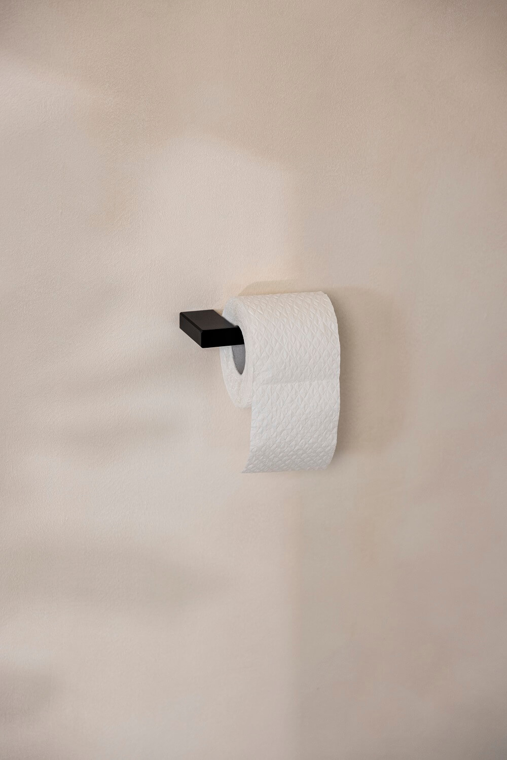 Eine Rolle Toilettenpapier auf einem modernen schwarzen Halter vor einer beigen Wand, mit sanften Schatten, die eine minimalistische Ästhetik schaffen.