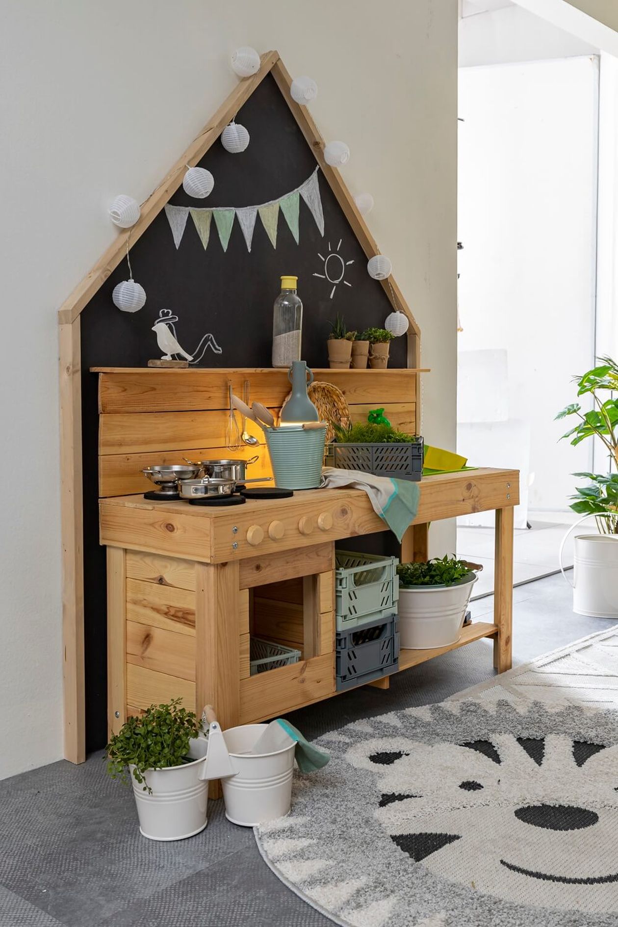 Naturbelassene Matschküche aus Holz mit vielen Pflanzen, kleinen Dekoartikeln und Kreidetafel im Hintergrund – Indoor-Spielküchen-Bereich.