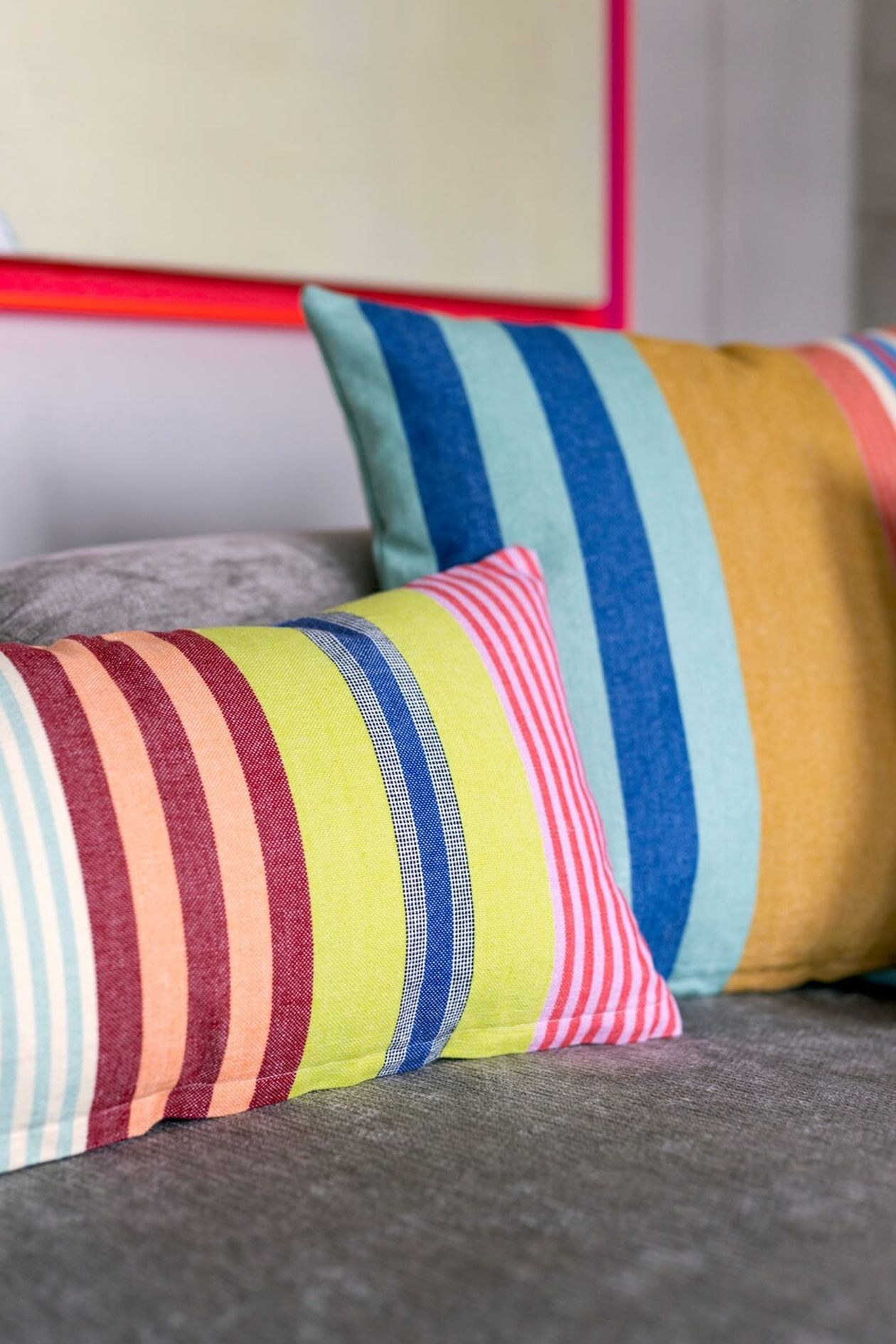 Zwei farbenfroh gestreifte Kissen – eines rechteckig, eines quadratisch – liegen auf einem grauen Sofa.
