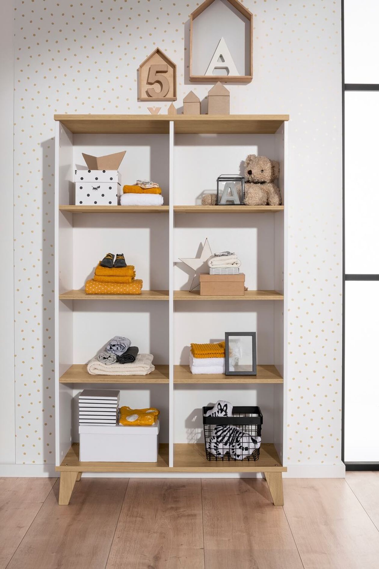 Weißes Standregal mit Holzdekor und offenen Fächern. Das Regal ist mit Kisten, Handtüchern, Stofftieren und Dekorationsartikeln bestückt, dahinter gepunktete Tapete.