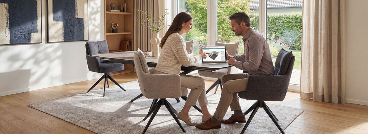 Ein Paar sitzt in einem hellen Esszimmer am Tisch und betrachtet etwas auf einem Laptop. Der Raum hat große Fenster und moderne Dekoration.