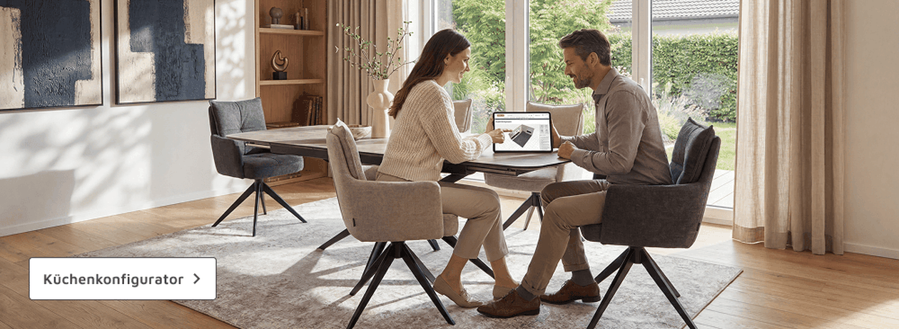 Ein Paar sitzt am Esstisch in einem modernen Wohnzimmer, schaut auf einen Laptop. Ein Button mit "Küchenkonfigurator" ist sichtbar.