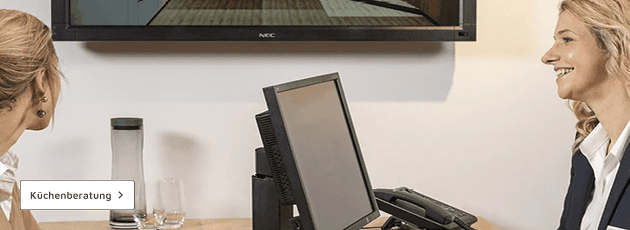 Zwei Frauen sitzen an einem Schreibtisch im Gespräch über Küchenplanung, ein Monitor und ein Telefon sind zu sehen.