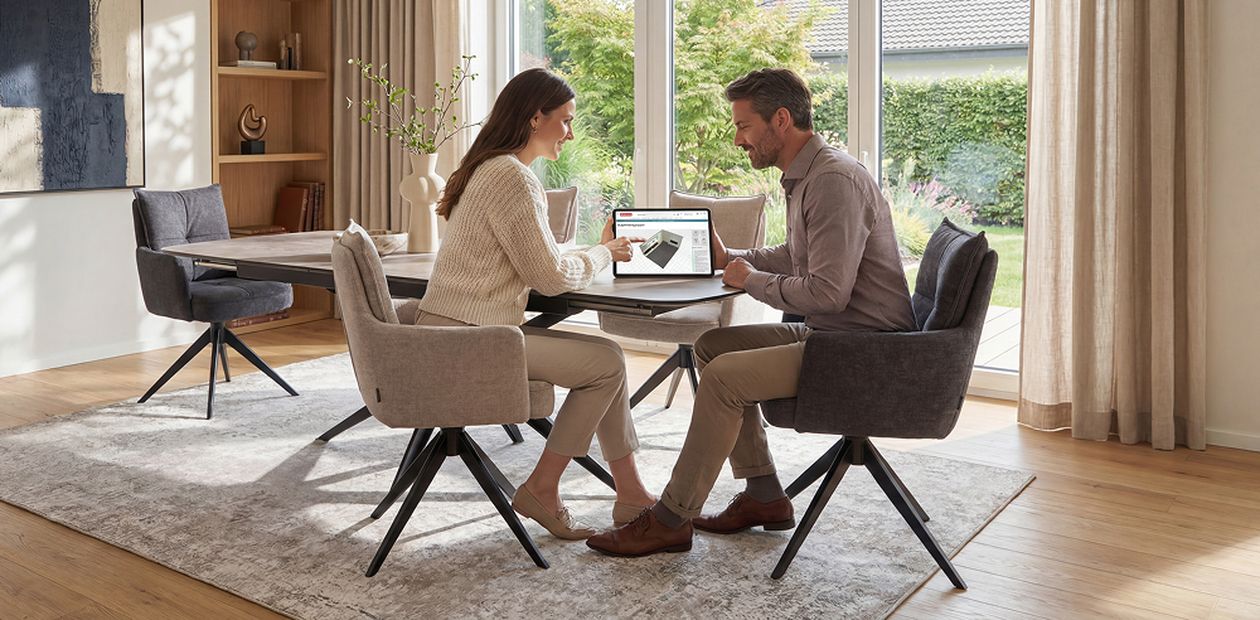 Ein Mann und eine Frau sitzen an einem Tisch und schauen gemeinsam auf einen Laptop. Das Zimmer ist modern eingerichtet mit großen Fenstern.