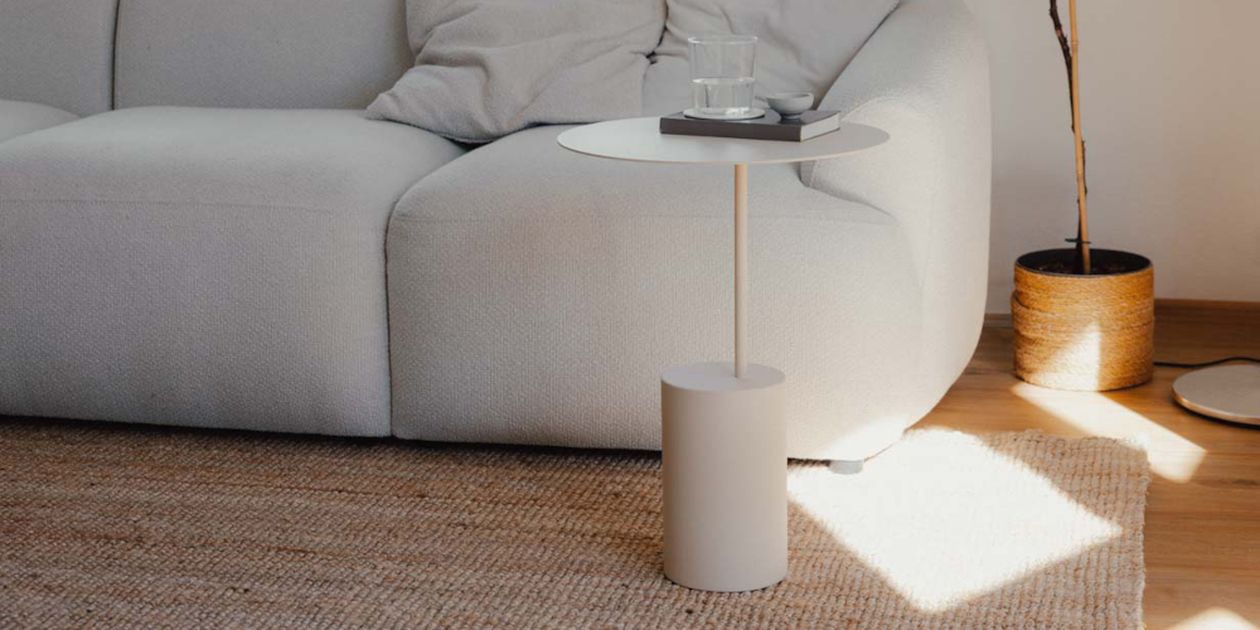 Minimalistisches Wohnzimmer mit einem weißen Sofa, einem runden Beistelltisch mit einem Glas und einem Buch darauf und einem gewebten Teppich auf einem Holzboden.