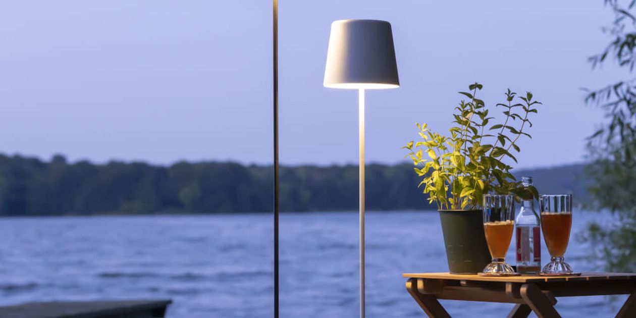 Eine Szene am See in der Abenddämmerung mit einer brennenden Lampe, einer Topfpflanze und Getränken auf einem Holztisch, mit Blick auf das ruhige Wasser und das von Bäumen gesäumte Ufer.