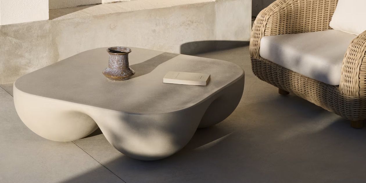 Eine moderne Außenumgebung mit einem skulpturalen Couchtisch, einem Keramikbecher, einem Buch und einem Korbsessel mit weißen Kissen auf einer sonnigen Terrasse.