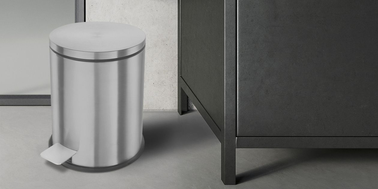 Edelstahl-Treteimer auf grauem Boden neben einem schwarzen Schrank vor hellgrauer Wand.