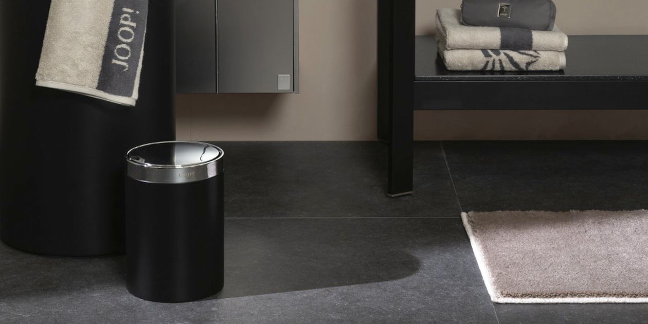 Modernes Badezimmer mit einem schwarzen Mülleimer, gefalteten Handtüchern auf einer Bank, einem beigefarbenen Handtuch, das über einer Stange hängt, und einem braunen Teppich auf einem dunklen Fliesenboden.