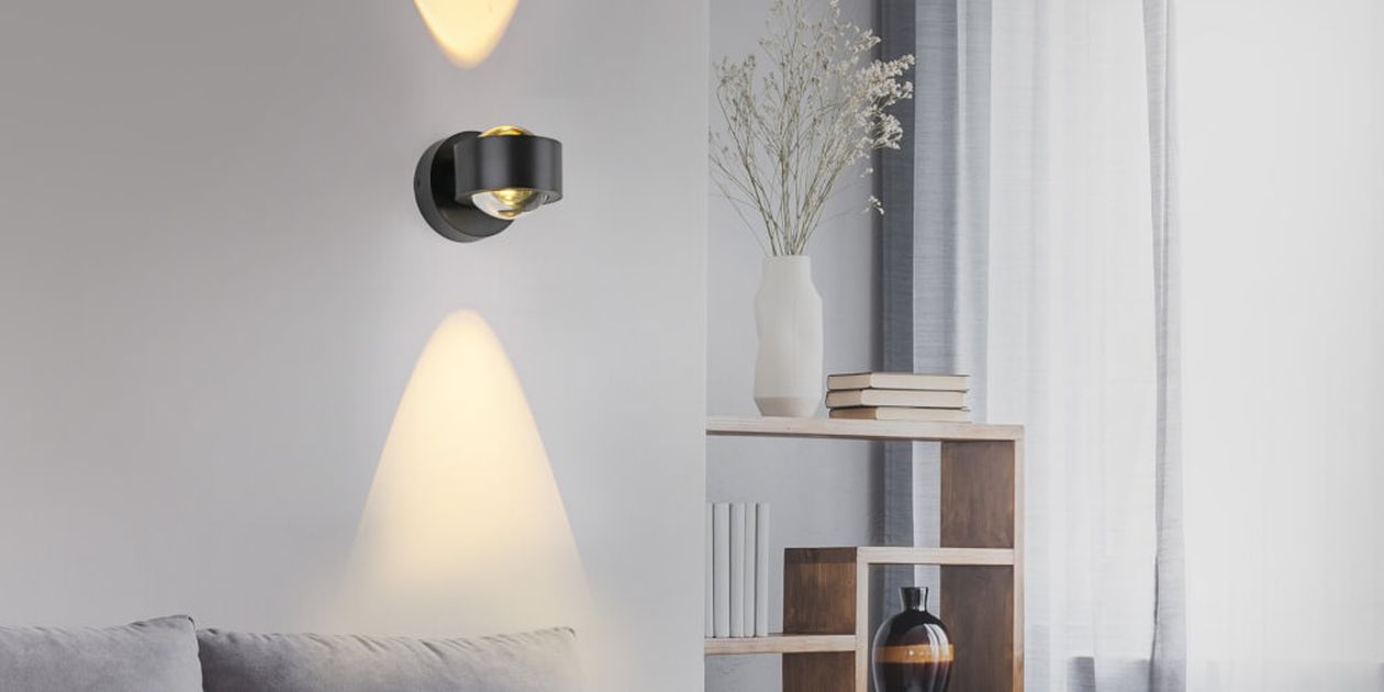 Modernes Wohnzimmer mit einer Wandleuchte, die ein sanftes Licht spendet, einem grauen Sofa und einem Holzregal mit Büchern, einer Vase und Dekoartikeln.