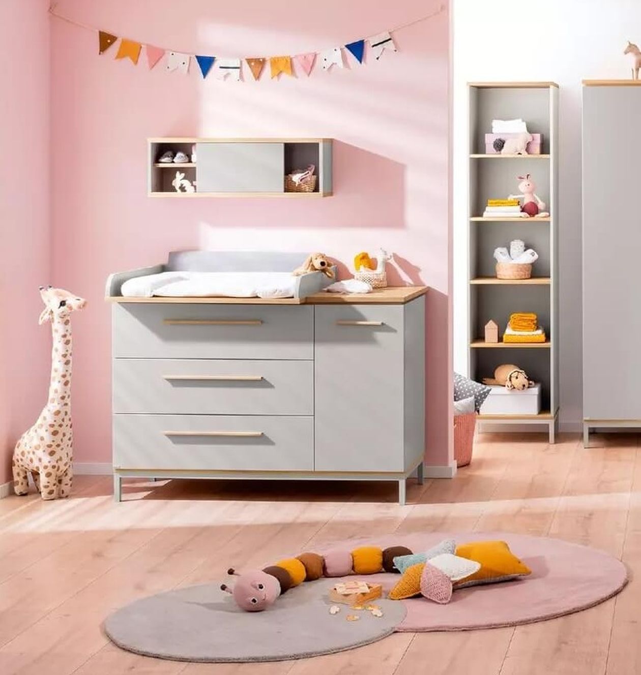 Helles Babyzimmer mit grau-holzfarbenem Möbelset bestehend aus Wickelkommode mit Aufsatz, Schrank und Babybett; im Hintergrund eine rosafarbene Wand und ein Giraffenkuscheltier.