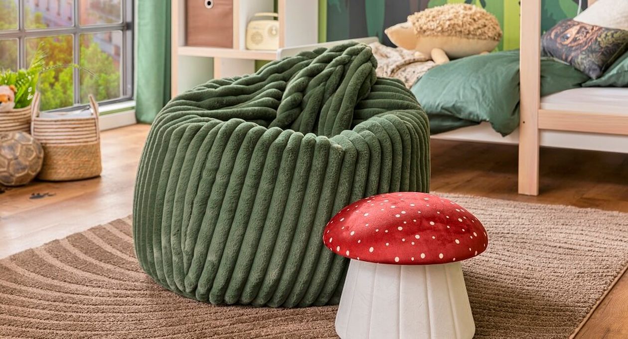 Ein gemütliches Zimmer mit einem grünen, strukturierten Sitzsack auf einem braunen Teppich und einem roten, pilzförmigen Hocker daneben. Im Hintergrund sind ein Bett und ein Fenster zu sehen.