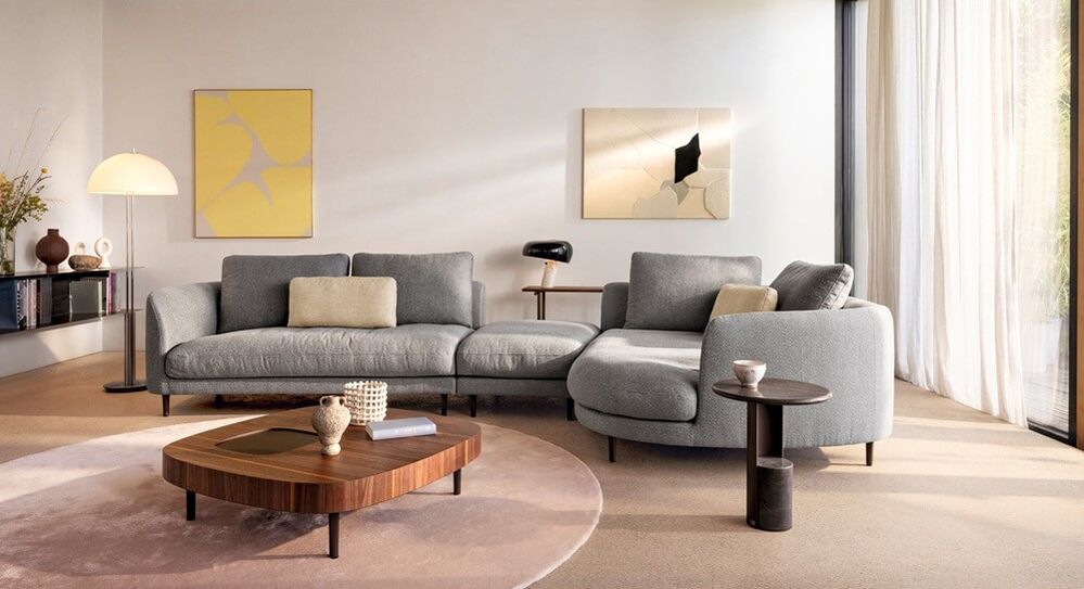 Elegantes graues Ecksofa mit organisch geschwungener Form in einem hellen Wohnzimmer mit Holzcouchtisch, rundem Teppich und modernen Kunstwerken an der Wand.
