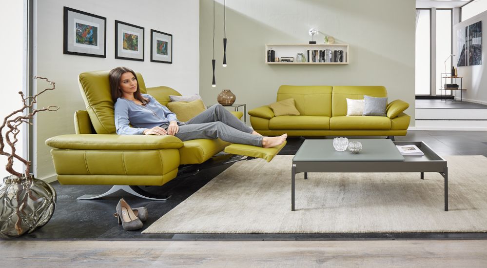 Zwei Sofas des Typs Vero aus den SEGMÜLLER WEKSTÄTTEN in grünem Leder stehen im rechten Winkel zueinander in einem modernen Wohnzimmer. Auf dem vorderen sitzt eine junge Frau in Relaxposition dank der ausgefahrenen Fußablage.