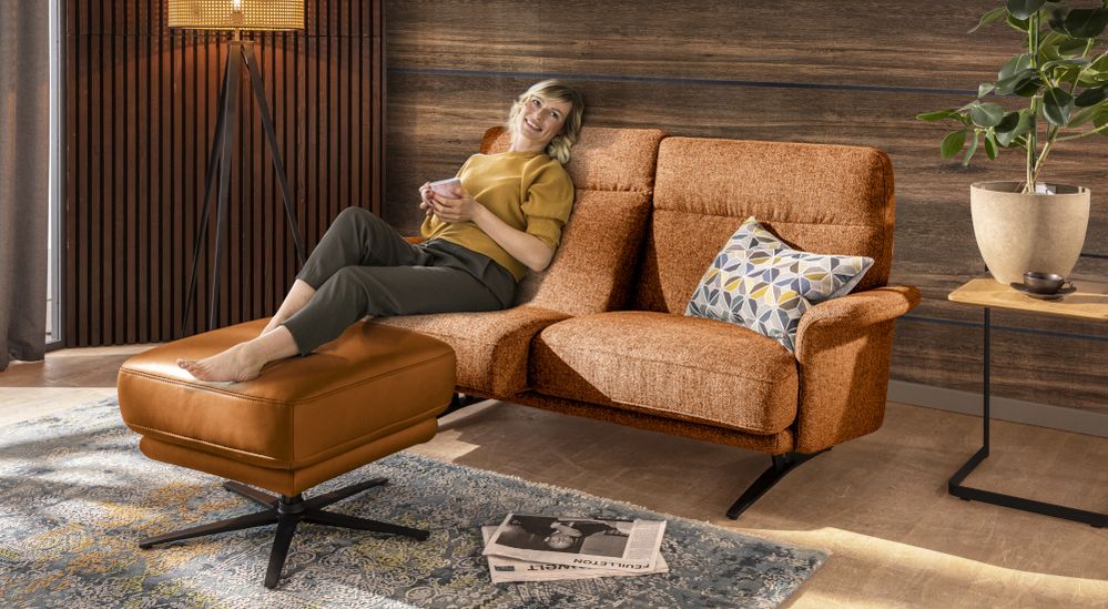EIne junge Frau liegt mit Teetasse auf dem orangenen Sofa Velino aus den SEGMÜLLER WERKSTÄTTEN. Ihre Füße liegen auf dem zugehörigen braunen Lederhocker.