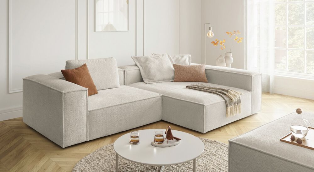 Modernes Wohnzimmer mit hellgrauem Ecksofa, braunen Kissen, einem runden weißen Couchtisch und sanftem natürlichem Licht durch ein großes Fenster.