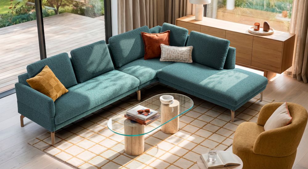 Grünes Ecksofa mit bunten Kissen, Glas-Couchtisch, gelbem Sessel und Holzmöbeln in einem modernen Wohnzimmer mit großen Fenstern.