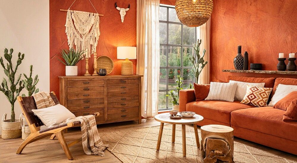 Gemütliches Wohnzimmer mit Terrakottawänden, orangefarbenem Sofa, Holzmöbeln, Makramee-Dekoration und üppigen Pflanzen. Ein großes Fenster lässt viel Tageslicht herein.