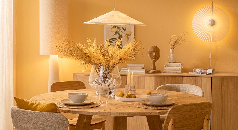 Gemütlicher Essbereich mit rundem Holztisch, gedeckt mit Geschirr und Kerzen. Gelbe Wände, warmes Licht, moderne Dekorationen.