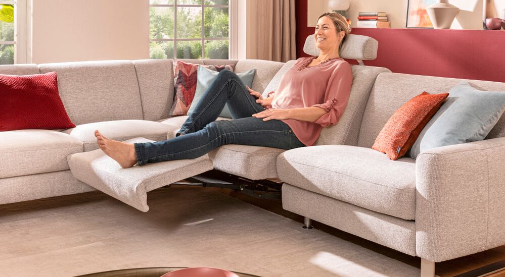 Frau entspannt sich auf einem hellgrauen Sofa mit zurückgelehnter Fußstütze, umgeben von bunten Kissen, in einem hellen Wohnzimmer.