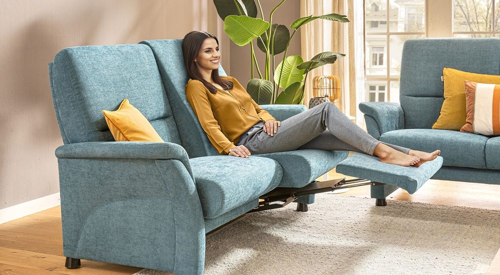 Frau entspannt auf einem blauen Liegesofa mit gelben Kissen in einem hellen Wohnzimmer mit Pflanzen und großen Fenstern.