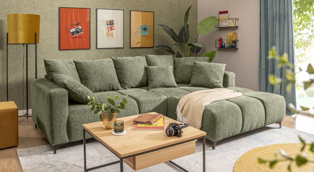 Grüne Eckcouch, Holztisch und bunte Wandbilder in hellem Wohnzimmer. Pflanzen und Stehlampe sorgen für Gemütlichkeit.