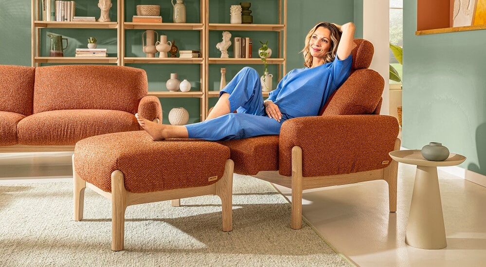 Eine Frau in blauem Outfit entspannt sich auf einem orangefarbenen Sessel mit passender Fußstütze in einem stilvollen Wohnzimmer mit Regalen und Dekoration.