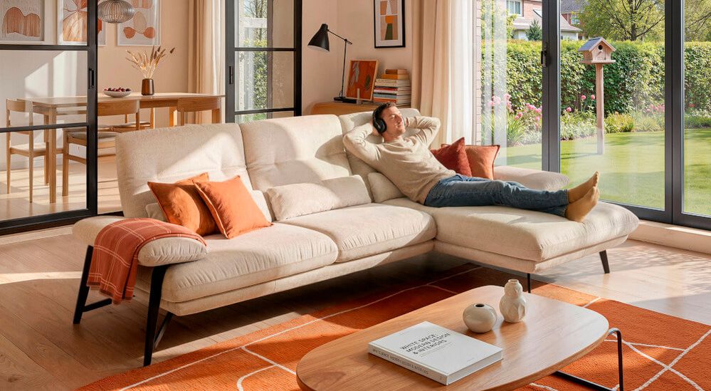 Eine Person entspannt sich auf einem modernen cremefarbenen Sofa mit orangefarbenen Kissen und trägt Kopfhörer in einem sonnendurchfluteten Wohnzimmer mit Blick in den Garten.