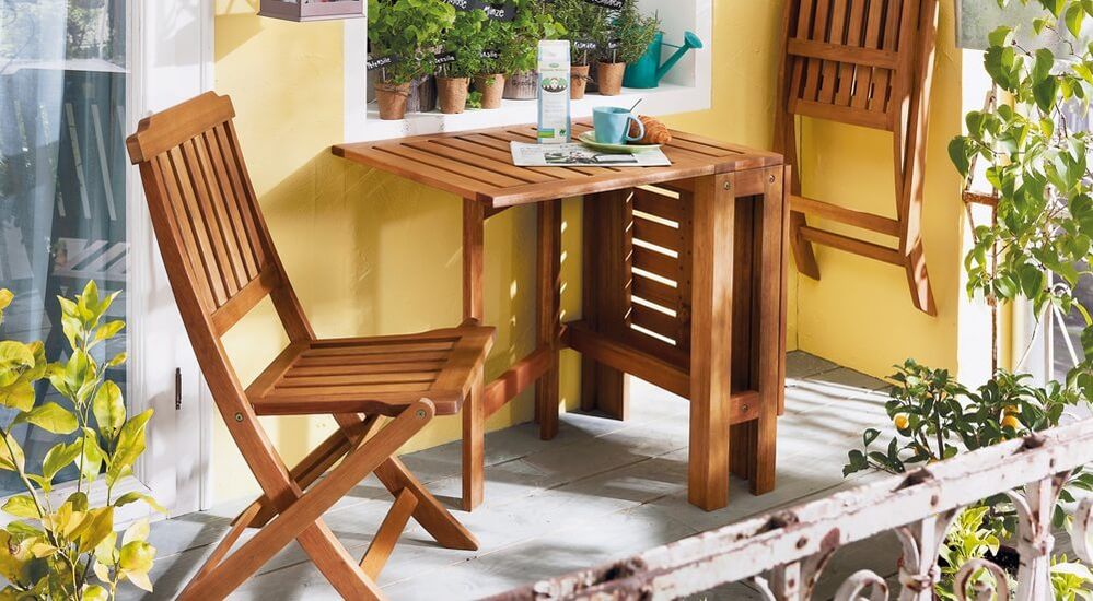 Gemütlicher Balkon mit Holztisch und Stühlen, Topfpflanzen, einer Zeitschrift und einer Tasse Kaffee an einem sonnigen Tag.