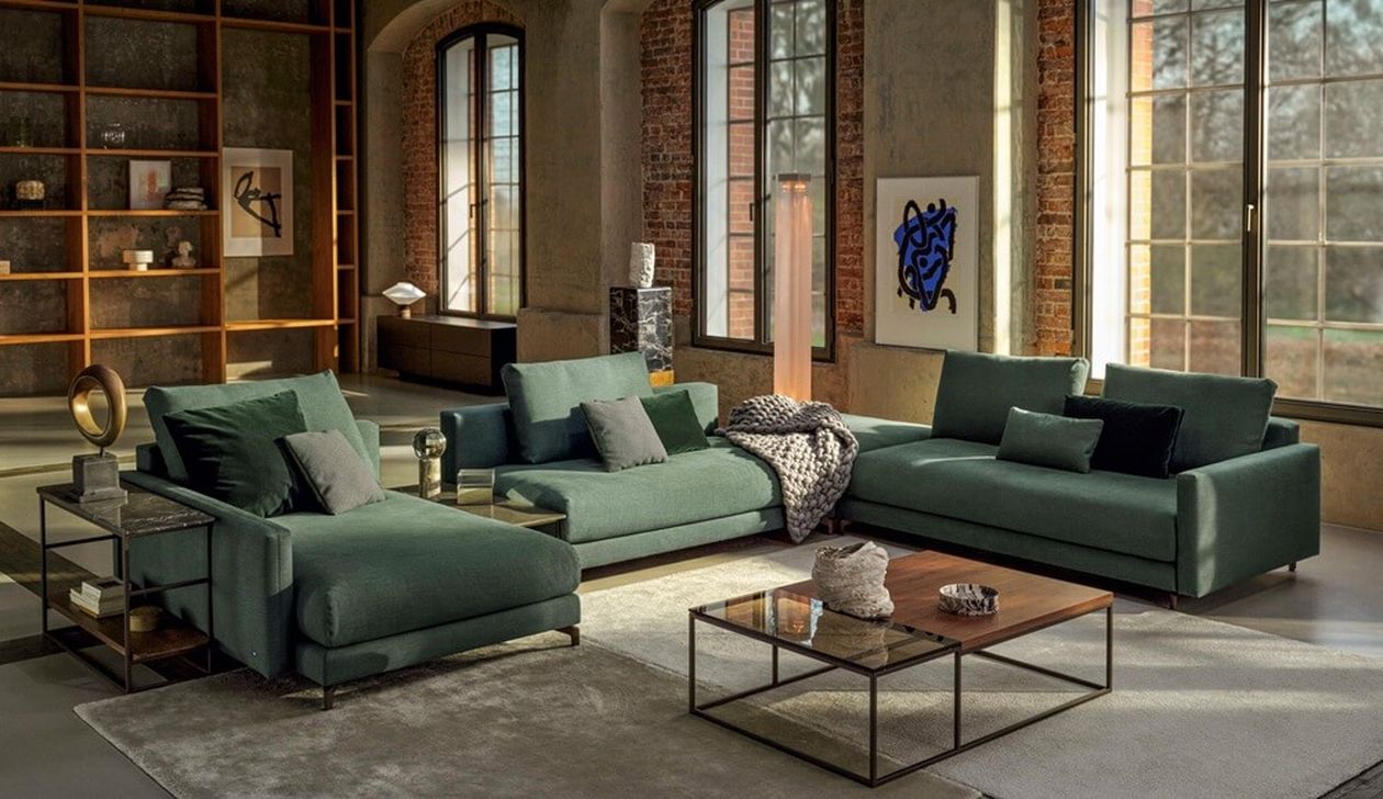 Großzügige grüne Wohnlandschaft in einem Loft mit hohen Fenstern, Backstein-Details, Regalen und einem modernen Couchtisch vor dem Sofa.