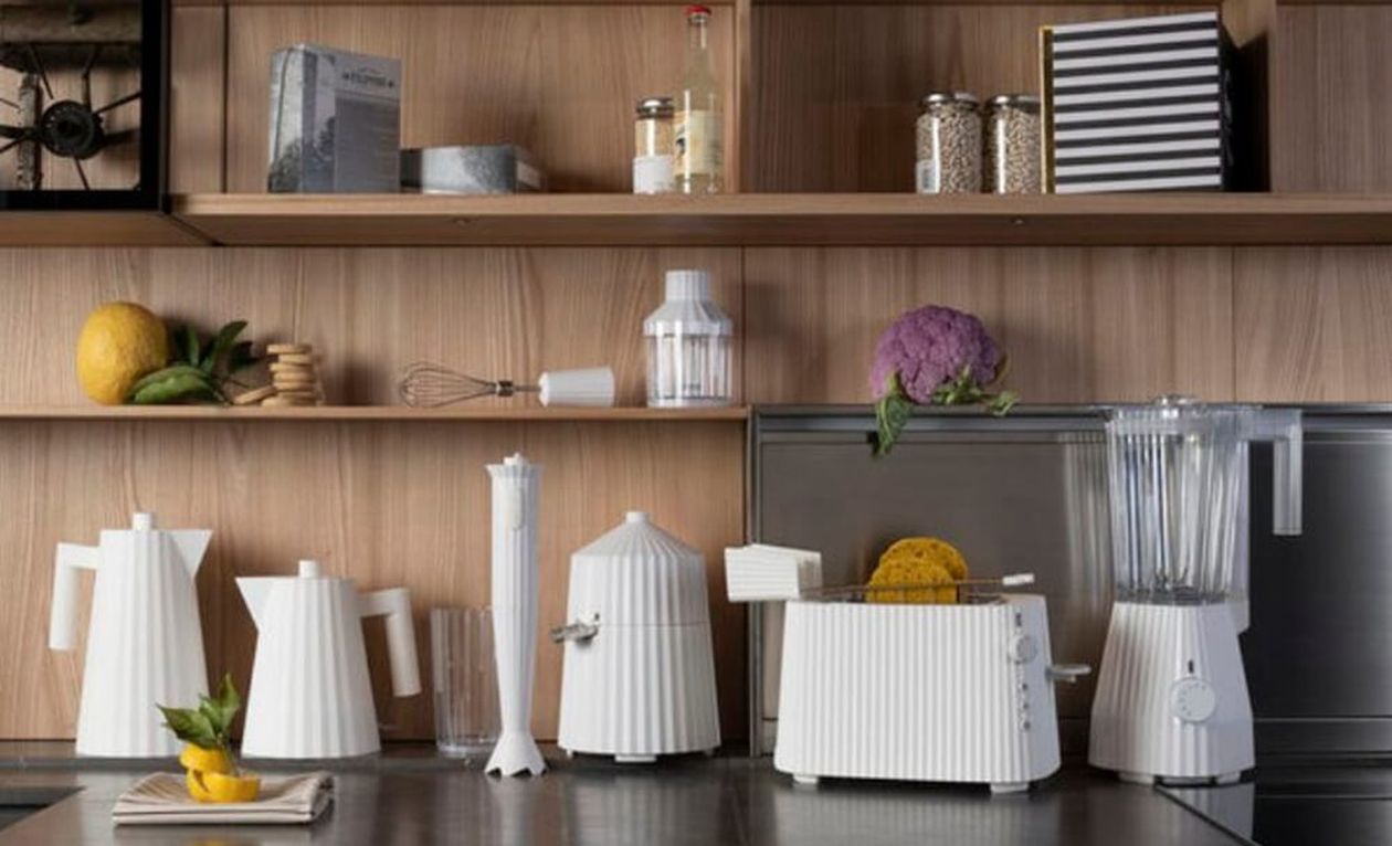 Weiße, modern designte Küchengeräte von Alessi, darunter ein Toaster, stehen dekorativ auf einer Edelstahl-Arbeitsplatte vor einer Holzwand. Im Hintergrund befinden sich offene Regale mit Küchenutensilien und Lebensmitteln.