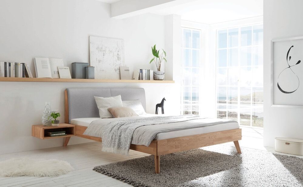Minimalistisches Schlafzimmer mit einem Holzbett, grauer Bettwäsche, einem schwebenden Regal mit Büchern, abstrakter Kunst, einem großen Fenster und einem gemütlichen grauen Teppich.