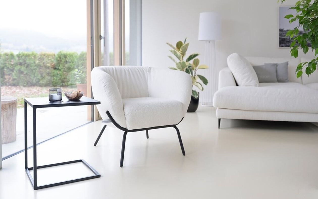 Weißer Polstersessel mit Bouclé-Bezug und filigranen schwarzen Metallbeinen in einem hellen, modernen Wohnzimmer. Daneben ein schwarzer Beistelltisch mit Dekoobjekten. Im Hintergrund ein weißes Sofa, eine weiße Stehleuchte, Zimmerpflanzen und eine große Glasfront mit Blick ins Grüne.