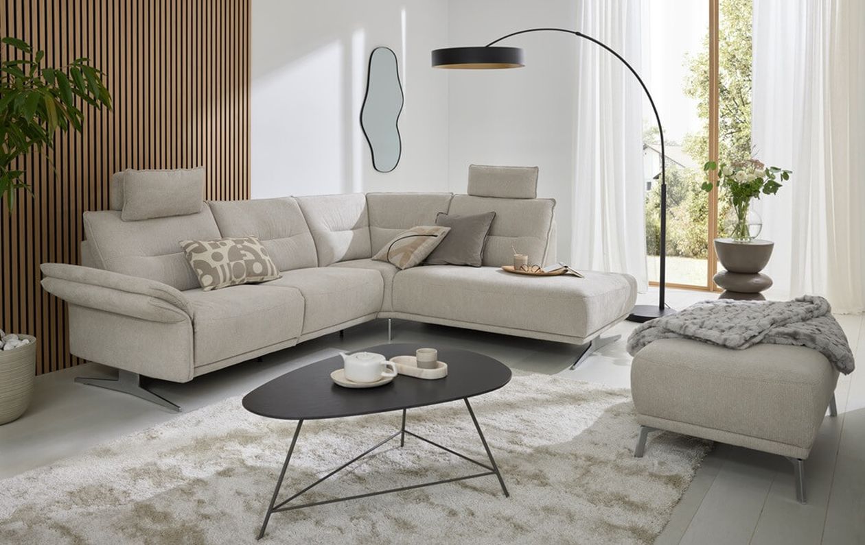 Modernes Wohnzimmer mit beigem Ecksofa, schwarzem ovalem Couchtisch mit filigranem Metallgestell, passender Bogenleuchte und Hocker auf einem weißen Hochflorteppich.