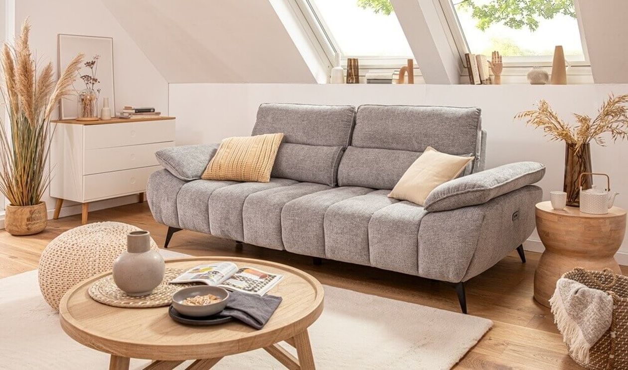 Graues Dreisitzer-Sofa mit verstellbaren Kopfstützen und gelben Dekokissen in einem hellen Dachgeschoss-Wohnzimmer mit Holzboden und natürlicher Dekoration.