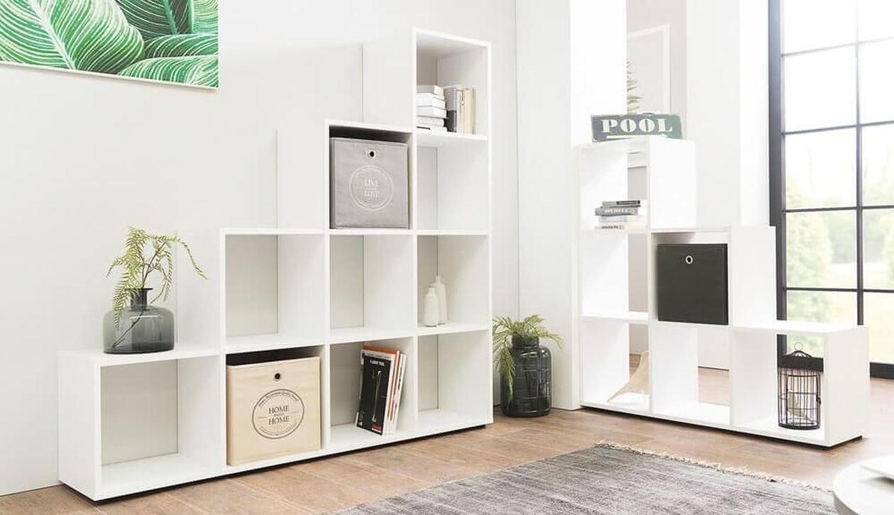 Zwei weiße Stufenregale in Treppenform stehen vor einer hellen Wand mit großem Fenster. Die offenen Fächer sind mit Büchern, Pflanzen und Aufbewahrungsboxen in Grau, Schwarz und Holzoptik dekoriert. Darüber ein großes Bild mit grünen Blättern.