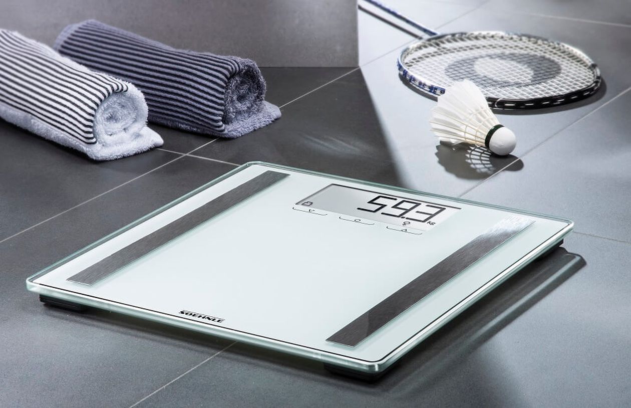Digitale Personenwaage auf gefliestem Boden, die 59,3 kg anzeigt. In der Nähe liegen gerollte Handtücher, ein Badmintonschläger und ein Federball.