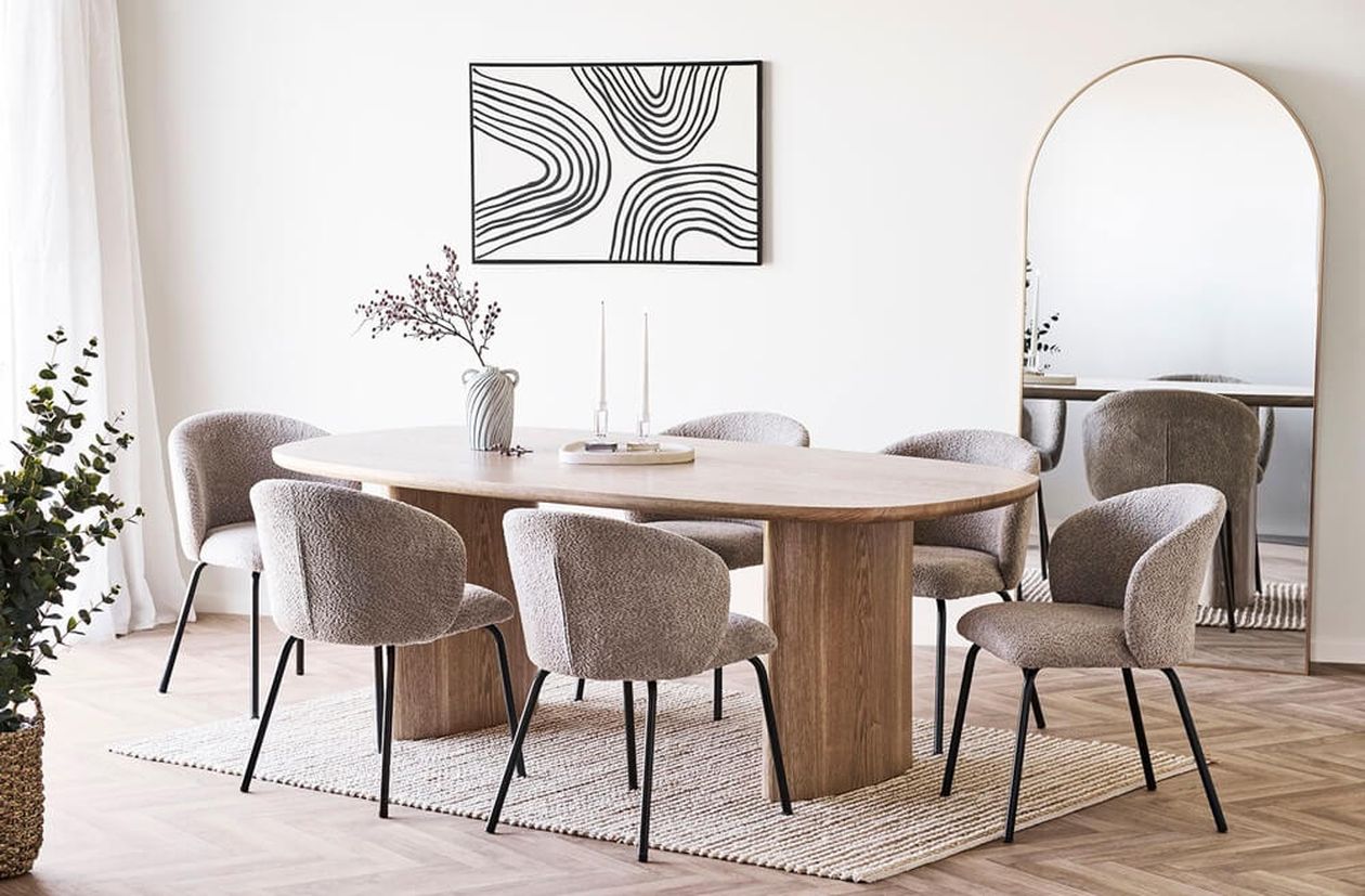 Ovaler Esstisch aus hellem Holz mit massivem Mittelfuß, umgeben von sechs taupefarbenen Bouclé-Stühlen in einem modernen Esszimmer mit Wandbild und Standspiegel.