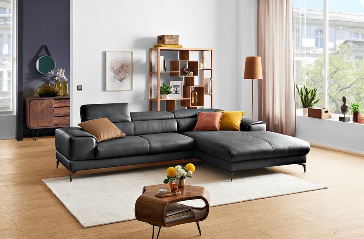Modernes Wohnzimmer mit schwarzem Sofa, bunten Kissen, Couchtisch aus Holz, Bücherregal und großen Fenstern mit Tageslicht.