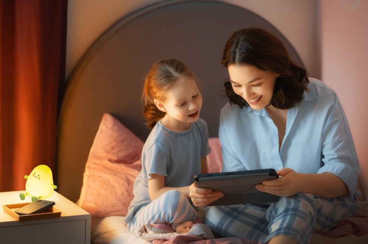 Mutter und Tochter sitzen abends auf einem Bett und schauen gemeinsam auf ein Tablet. Auf dem Nachttisch leuchtet eine kleine Akku-Nachtleuchte in Form einer grünen Avocado mit freundlichem Gesicht und sorgt für sanftes, warmes Licht im Kinderzimmer.
